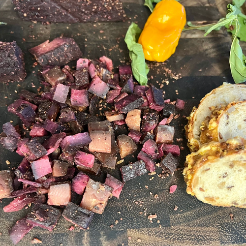 Diced Anna Meru Alpine Smoked Vegan Cubes on a dark wooden board, served alongside sliced crusty bread, fresh basil, and a yellow pepper.