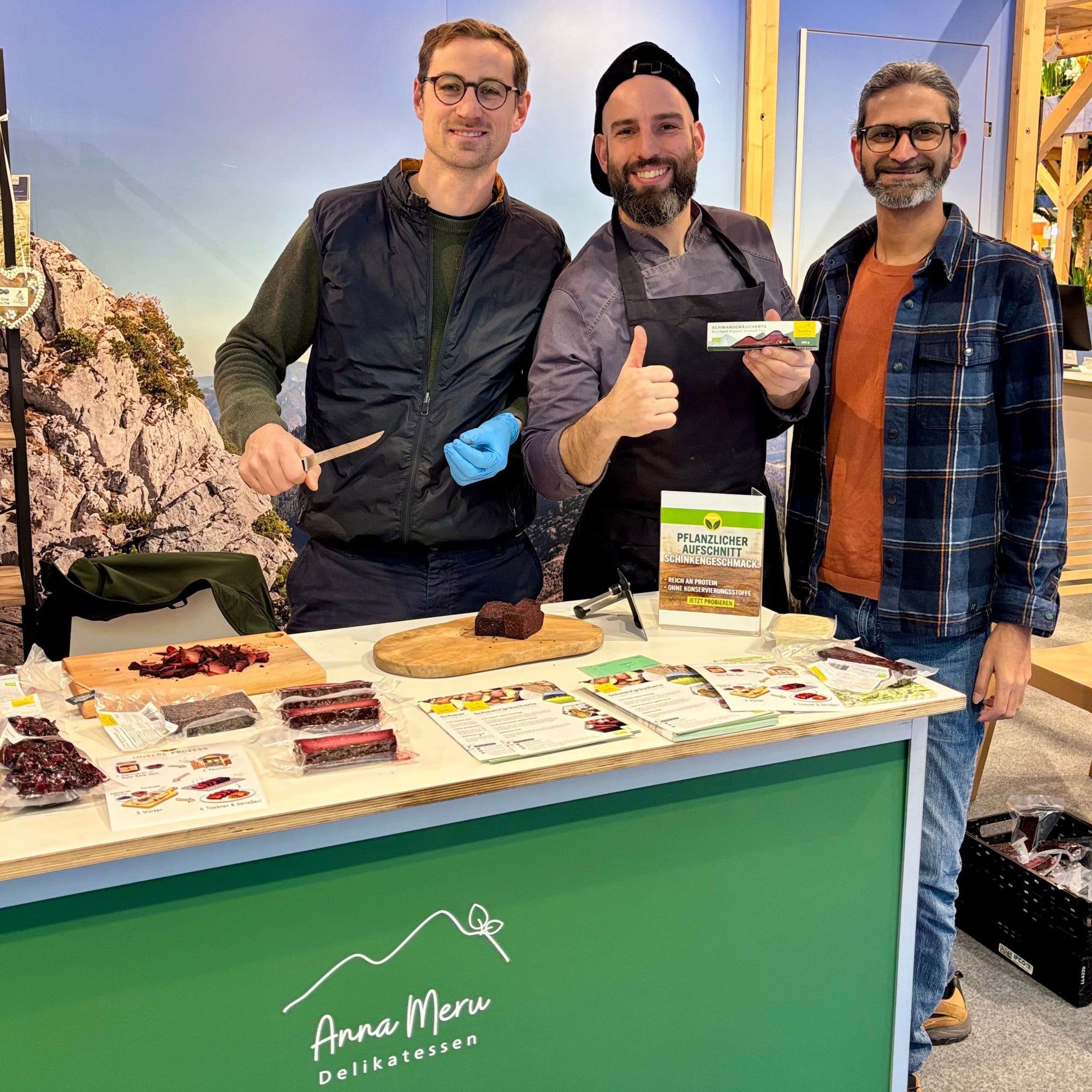 The three founders of Anna Meru smiling behind a market stand, showcasing their artisanal plant-based products.Moritz-Patrick-Ranjan